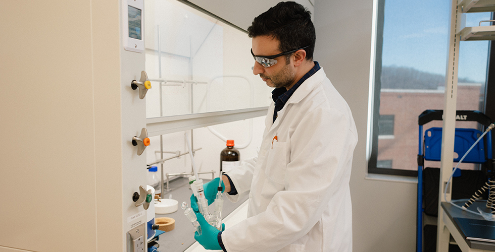 Photo of male student wearing safety glasses and white lab coat holding glass tubes.