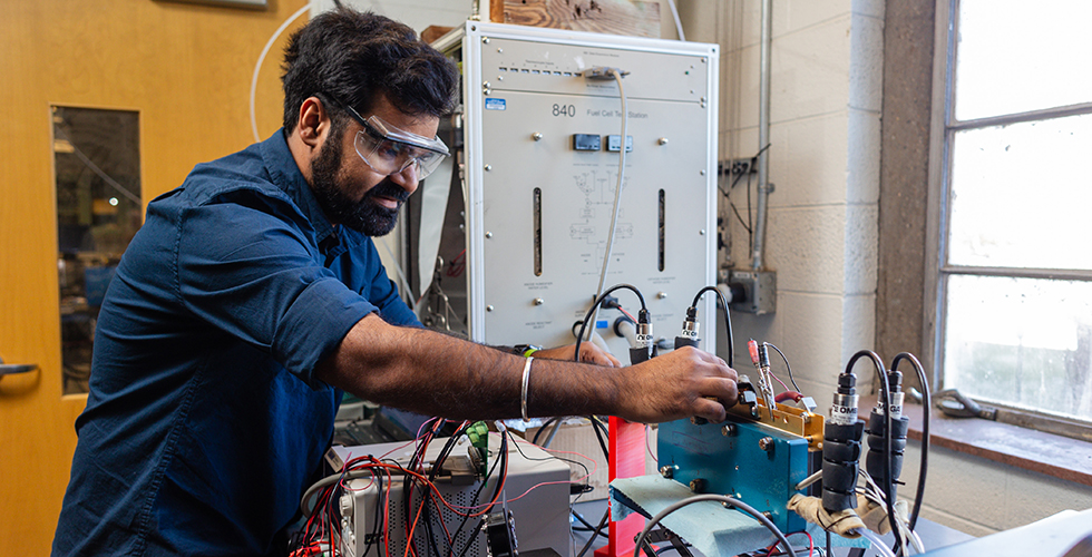 Photo of male student with dark hair wearing blue shirt and safety glasses working on fuel cell.