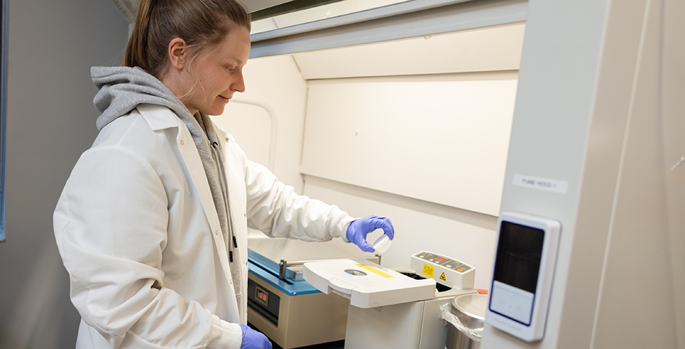 Photo of female student wearing white lab coat holding sample under a lab hood.